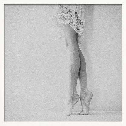 A framed, grainy black and white photograph of a dancer's legs and feet. The dancer stands on tiptoes against a white wall, wearing a delicate lace skirt. Poster