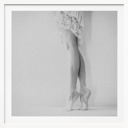A framed, grainy black and white photograph of a dancer's legs and feet. The dancer stands on tiptoes against a white wall, wearing a delicate lace skirt. Poster