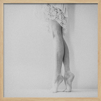 A framed, grainy black and white photograph of a dancer's legs and feet. The dancer stands on tiptoes against a white wall, wearing a delicate lace skirt. Poster