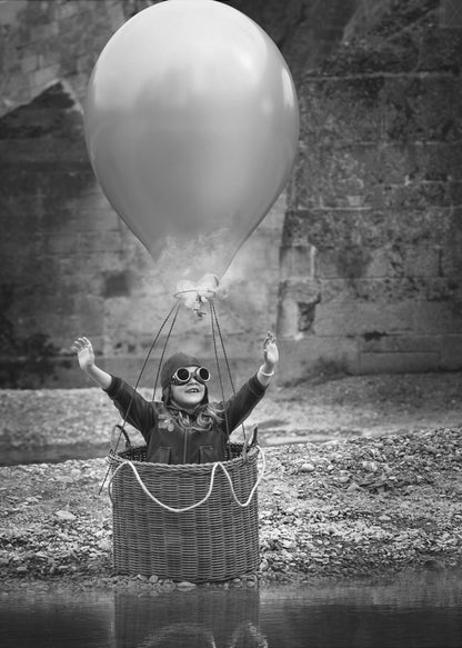 A whimsical black and white photograph of a young girl wearing an aviator cap and goggles, joyfully raising her arms in the air while standing in a wicker basket attached to a large balloon, mimicking a hot air balloon. The scene is set by the water with a stone wall in the background. Poster