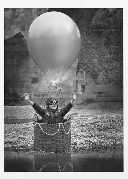 A whimsical black and white photograph of a young girl wearing an aviator cap and goggles, joyfully raising her arms in the air while standing in a wicker basket attached to a large balloon, mimicking a hot air balloon. The scene is set by the water with a stone wall in the background. Poster
