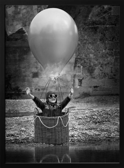 A whimsical black and white photograph of a young girl wearing an aviator cap and goggles, joyfully raising her arms in the air while standing in a wicker basket attached to a large balloon, mimicking a hot air balloon. The scene is set by the water with a stone wall in the background. Poster