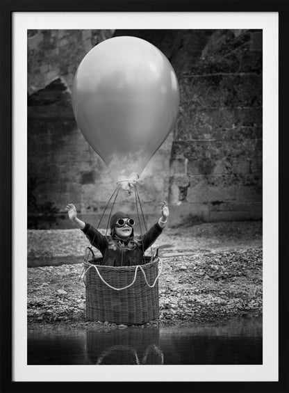 A whimsical black and white photograph of a young girl wearing an aviator cap and goggles, joyfully raising her arms in the air while standing in a wicker basket attached to a large balloon, mimicking a hot air balloon. The scene is set by the water with a stone wall in the background. Poster