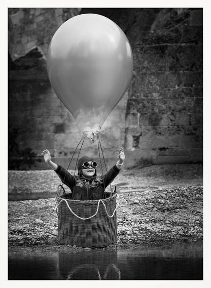 A whimsical black and white photograph of a young girl wearing an aviator cap and goggles, joyfully raising her arms in the air while standing in a wicker basket attached to a large balloon, mimicking a hot air balloon. The scene is set by the water with a stone wall in the background. Poster