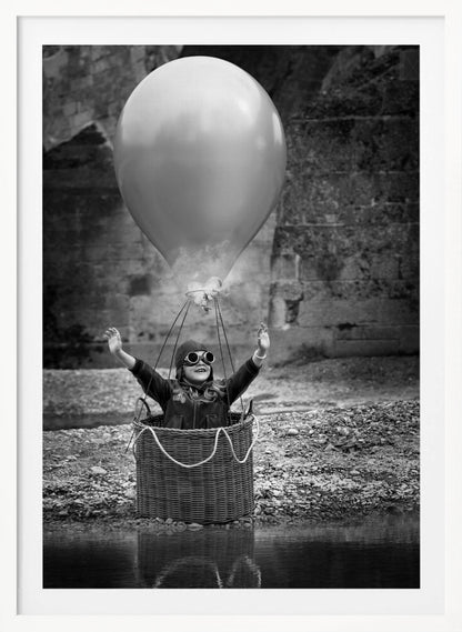 A whimsical black and white photograph of a young girl wearing an aviator cap and goggles, joyfully raising her arms in the air while standing in a wicker basket attached to a large balloon, mimicking a hot air balloon. The scene is set by the water with a stone wall in the background. Poster