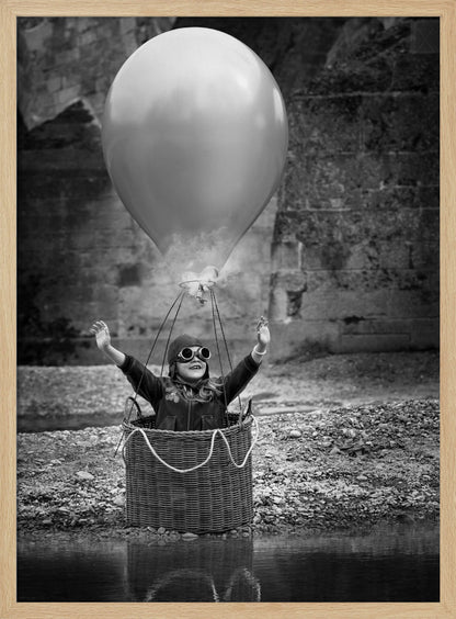 A whimsical black and white photograph of a young girl wearing an aviator cap and goggles, joyfully raising her arms in the air while standing in a wicker basket attached to a large balloon, mimicking a hot air balloon. The scene is set by the water with a stone wall in the background. Poster