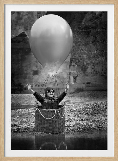 A whimsical black and white photograph of a young girl wearing an aviator cap and goggles, joyfully raising her arms in the air while standing in a wicker basket attached to a large balloon, mimicking a hot air balloon. The scene is set by the water with a stone wall in the background. Poster
