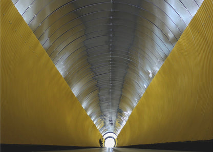 A symmetrical, perspective view down a modern, circular tunnel. The lower walls are bright yellow, creating two diagonal lines that converge towards a bright, white light at the far end. The arched ceiling is made of reflective silver metal panels, and a small silhouette stands near the exit. Artwork
