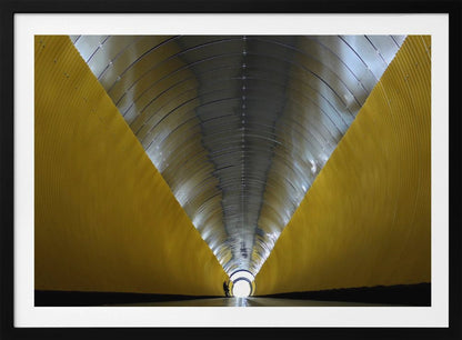 A symmetrical, perspective view down a modern, circular tunnel. The lower walls are bright yellow, creating two diagonal lines that converge towards a bright, white light at the far end. The arched ceiling is made of reflective silver metal panels, and a small silhouette stands near the exit. Artwork