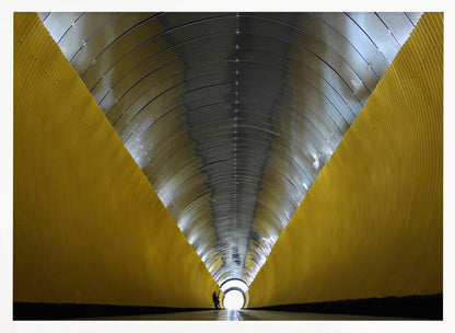 A symmetrical, perspective view down a modern, circular tunnel. The lower walls are bright yellow, creating two diagonal lines that converge towards a bright, white light at the far end. The arched ceiling is made of reflective silver metal panels, and a small silhouette stands near the exit. Artwork