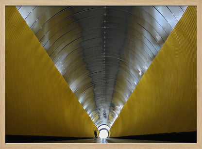 A symmetrical, perspective view down a modern, circular tunnel. The lower walls are bright yellow, creating two diagonal lines that converge towards a bright, white light at the far end. The arched ceiling is made of reflective silver metal panels, and a small silhouette stands near the exit. Artwork