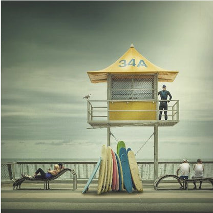 A vintage-style photograph of a yellow lifeguard tower labeled '34A' on a beach boardwalk under an overcast, greenish sky. A lifeguard in a wetsuit stands on the balcony, while a collection of colorful surfboards leans against the base. People are seen relaxing on benches nearby. Print