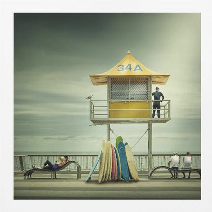 A vintage-style photograph of a yellow lifeguard tower labeled '34A' on a beach boardwalk under an overcast, greenish sky. A lifeguard in a wetsuit stands on the balcony, while a collection of colorful surfboards leans against the base. People are seen relaxing on benches nearby. Print