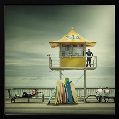 A vintage-style photograph of a yellow lifeguard tower labeled '34A' on a beach boardwalk under an overcast, greenish sky. A lifeguard in a wetsuit stands on the balcony, while a collection of colorful surfboards leans against the base. People are seen relaxing on benches nearby. Print