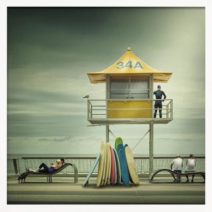 A vintage-style photograph of a yellow lifeguard tower labeled '34A' on a beach boardwalk under an overcast, greenish sky. A lifeguard in a wetsuit stands on the balcony, while a collection of colorful surfboards leans against the base. People are seen relaxing on benches nearby. Print