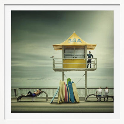 A vintage-style photograph of a yellow lifeguard tower labeled '34A' on a beach boardwalk under an overcast, greenish sky. A lifeguard in a wetsuit stands on the balcony, while a collection of colorful surfboards leans against the base. People are seen relaxing on benches nearby. Print