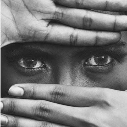 A powerful black and white close-up photograph of a person's face, framed in light wood. Their hands cover most of their face, one over the forehead and the other over the mouth, revealing only their intense, striking eyes that gaze directly forward. Print