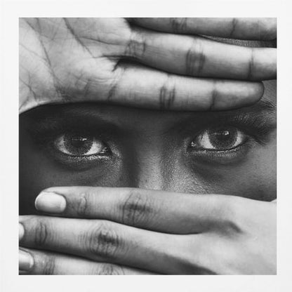 A powerful black and white close-up photograph of a person's face, framed in light wood. Their hands cover most of their face, one over the forehead and the other over the mouth, revealing only their intense, striking eyes that gaze directly forward. Print