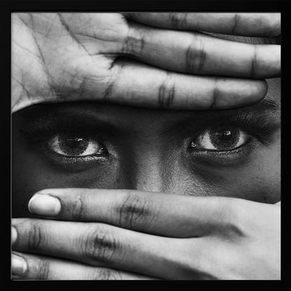 A powerful black and white close-up photograph of a person's face, framed in light wood. Their hands cover most of their face, one over the forehead and the other over the mouth, revealing only their intense, striking eyes that gaze directly forward. Print