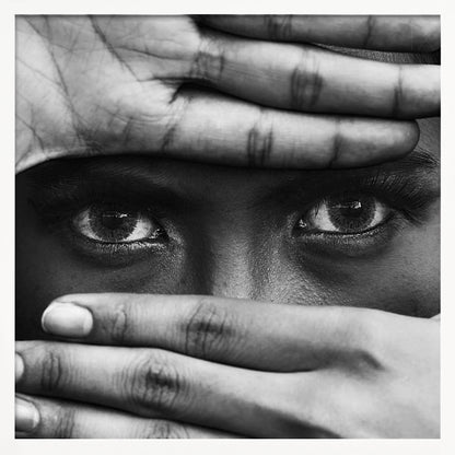 A powerful black and white close-up photograph of a person's face, framed in light wood. Their hands cover most of their face, one over the forehead and the other over the mouth, revealing only their intense, striking eyes that gaze directly forward. Print