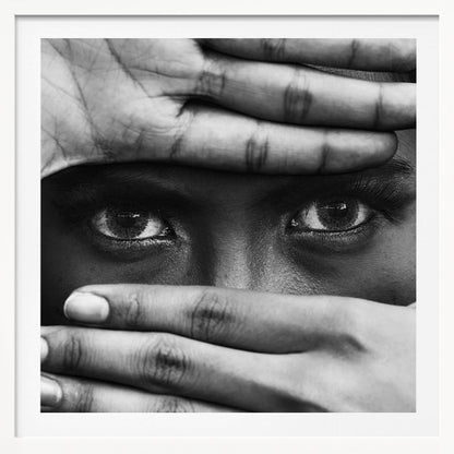 A powerful black and white close-up photograph of a person's face, framed in light wood. Their hands cover most of their face, one over the forehead and the other over the mouth, revealing only their intense, striking eyes that gaze directly forward. Print