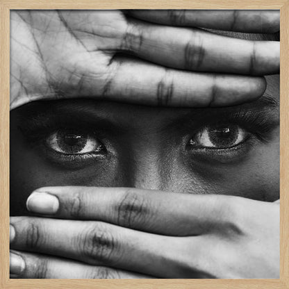 A powerful black and white close-up photograph of a person's face, framed in light wood. Their hands cover most of their face, one over the forehead and the other over the mouth, revealing only their intense, striking eyes that gaze directly forward. Print