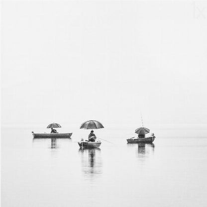 A minimalist black and white photograph of three fishermen in small boats on a calm, misty lake, each holding an umbrella. The image is framed in light wood. Artwork