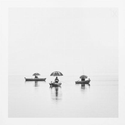 A minimalist black and white photograph of three fishermen in small boats on a calm, misty lake, each holding an umbrella. The image is framed in light wood. Artwork