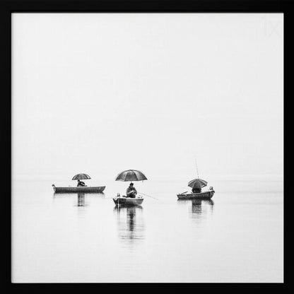 A minimalist black and white photograph of three fishermen in small boats on a calm, misty lake, each holding an umbrella. The image is framed in light wood. Artwork