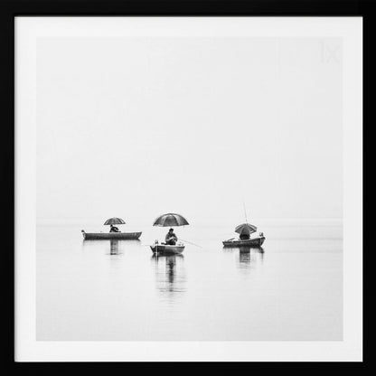 A minimalist black and white photograph of three fishermen in small boats on a calm, misty lake, each holding an umbrella. The image is framed in light wood. Artwork