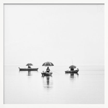 A minimalist black and white photograph of three fishermen in small boats on a calm, misty lake, each holding an umbrella. The image is framed in light wood. Artwork