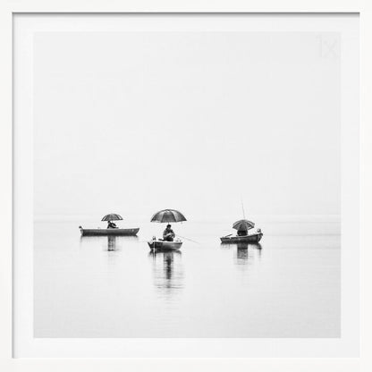 A minimalist black and white photograph of three fishermen in small boats on a calm, misty lake, each holding an umbrella. The image is framed in light wood. Artwork