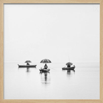 A minimalist black and white photograph of three fishermen in small boats on a calm, misty lake, each holding an umbrella. The image is framed in light wood. Artwork