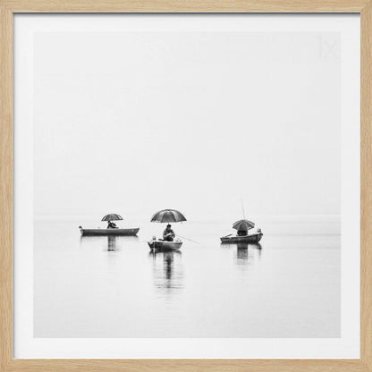 A minimalist black and white photograph of three fishermen in small boats on a calm, misty lake, each holding an umbrella. The image is framed in light wood. Artwork