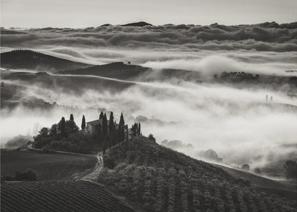A framed, black and white photograph of a dramatic Tuscan landscape. A villa surrounded by cypress trees sits on a foreground hill, while thick fog blankets the rolling hills and valleys below. In the distance, a sea of clouds stretches towards a mountain range. Wall Art