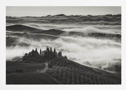 A framed, black and white photograph of a dramatic Tuscan landscape. A villa surrounded by cypress trees sits on a foreground hill, while thick fog blankets the rolling hills and valleys below. In the distance, a sea of clouds stretches towards a mountain range. Wall Art