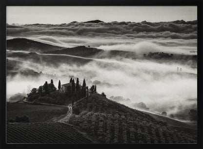 A framed, black and white photograph of a dramatic Tuscan landscape. A villa surrounded by cypress trees sits on a foreground hill, while thick fog blankets the rolling hills and valleys below. In the distance, a sea of clouds stretches towards a mountain range. Wall Art
