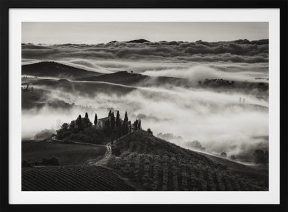 A framed, black and white photograph of a dramatic Tuscan landscape. A villa surrounded by cypress trees sits on a foreground hill, while thick fog blankets the rolling hills and valleys below. In the distance, a sea of clouds stretches towards a mountain range. Wall Art