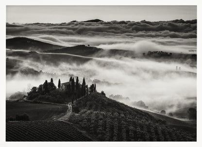 A framed, black and white photograph of a dramatic Tuscan landscape. A villa surrounded by cypress trees sits on a foreground hill, while thick fog blankets the rolling hills and valleys below. In the distance, a sea of clouds stretches towards a mountain range. Wall Art