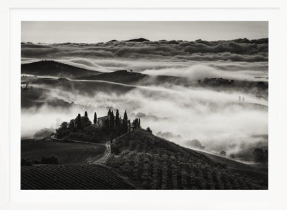 A framed, black and white photograph of a dramatic Tuscan landscape. A villa surrounded by cypress trees sits on a foreground hill, while thick fog blankets the rolling hills and valleys below. In the distance, a sea of clouds stretches towards a mountain range. Wall Art