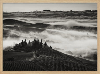 A framed, black and white photograph of a dramatic Tuscan landscape. A villa surrounded by cypress trees sits on a foreground hill, while thick fog blankets the rolling hills and valleys below. In the distance, a sea of clouds stretches towards a mountain range. Wall Art