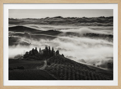 A framed, black and white photograph of a dramatic Tuscan landscape. A villa surrounded by cypress trees sits on a foreground hill, while thick fog blankets the rolling hills and valleys below. In the distance, a sea of clouds stretches towards a mountain range. Wall Art