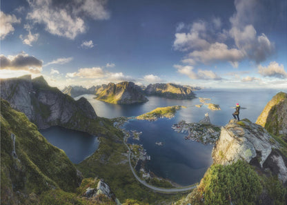A breathtaking panoramic photograph of the Lofoten Islands in Norway from a high vantage point. A person stands on a rocky cliff with arms outstretched, overlooking a stunning landscape of deep blue fjords, lush green mountains, and the small fishing village of Reine below. The sky is a vibrant blue with scattered white clouds. Artwork