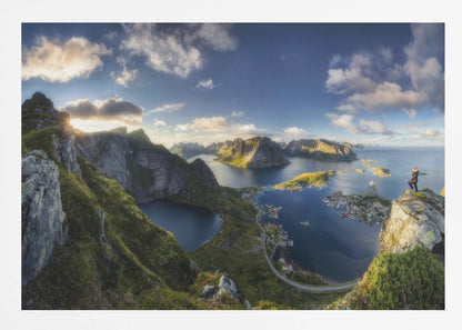 A breathtaking panoramic photograph of the Lofoten Islands in Norway from a high vantage point. A person stands on a rocky cliff with arms outstretched, overlooking a stunning landscape of deep blue fjords, lush green mountains, and the small fishing village of Reine below. The sky is a vibrant blue with scattered white clouds. Artwork