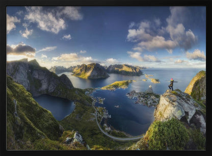 A breathtaking panoramic photograph of the Lofoten Islands in Norway from a high vantage point. A person stands on a rocky cliff with arms outstretched, overlooking a stunning landscape of deep blue fjords, lush green mountains, and the small fishing village of Reine below. The sky is a vibrant blue with scattered white clouds. Artwork