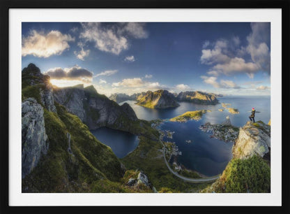 A breathtaking panoramic photograph of the Lofoten Islands in Norway from a high vantage point. A person stands on a rocky cliff with arms outstretched, overlooking a stunning landscape of deep blue fjords, lush green mountains, and the small fishing village of Reine below. The sky is a vibrant blue with scattered white clouds. Artwork