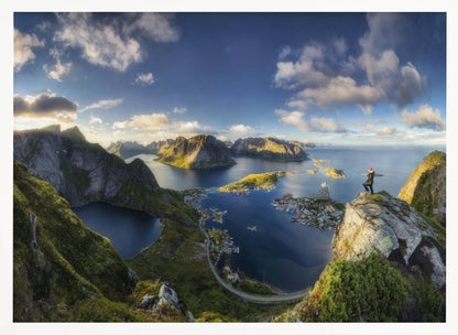 A breathtaking panoramic photograph of the Lofoten Islands in Norway from a high vantage point. A person stands on a rocky cliff with arms outstretched, overlooking a stunning landscape of deep blue fjords, lush green mountains, and the small fishing village of Reine below. The sky is a vibrant blue with scattered white clouds. Artwork