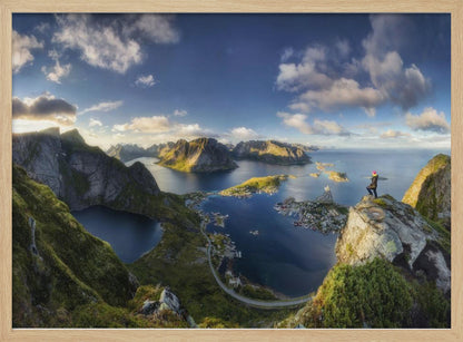 A breathtaking panoramic photograph of the Lofoten Islands in Norway from a high vantage point. A person stands on a rocky cliff with arms outstretched, overlooking a stunning landscape of deep blue fjords, lush green mountains, and the small fishing village of Reine below. The sky is a vibrant blue with scattered white clouds. Artwork