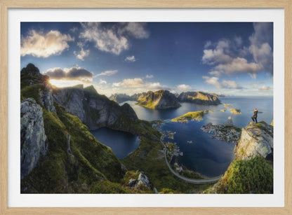 A breathtaking panoramic photograph of the Lofoten Islands in Norway from a high vantage point. A person stands on a rocky cliff with arms outstretched, overlooking a stunning landscape of deep blue fjords, lush green mountains, and the small fishing village of Reine below. The sky is a vibrant blue with scattered white clouds. Artwork