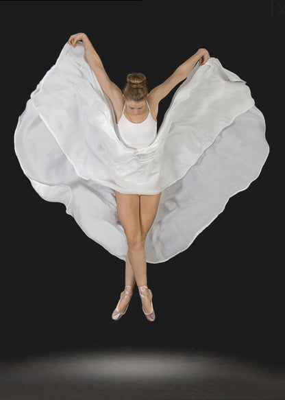 A high-angle studio photograph of a graceful ballet dancer suspended mid-air against a solid black background. She wears a white leotard and pointe shoes, with her legs crossed. Her arms are outstretched, holding a large, flowing white fabric that creates a shape like wings around her. Decor